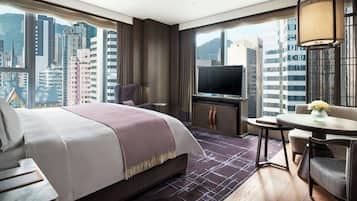A hotel room with a large bed, a television, a dining table, and a view of tall buildings.