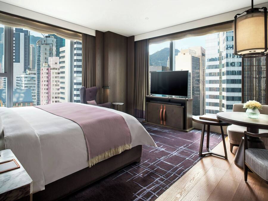 A hotel room with a large bed, a television, a dining table, and a view of tall buildings.