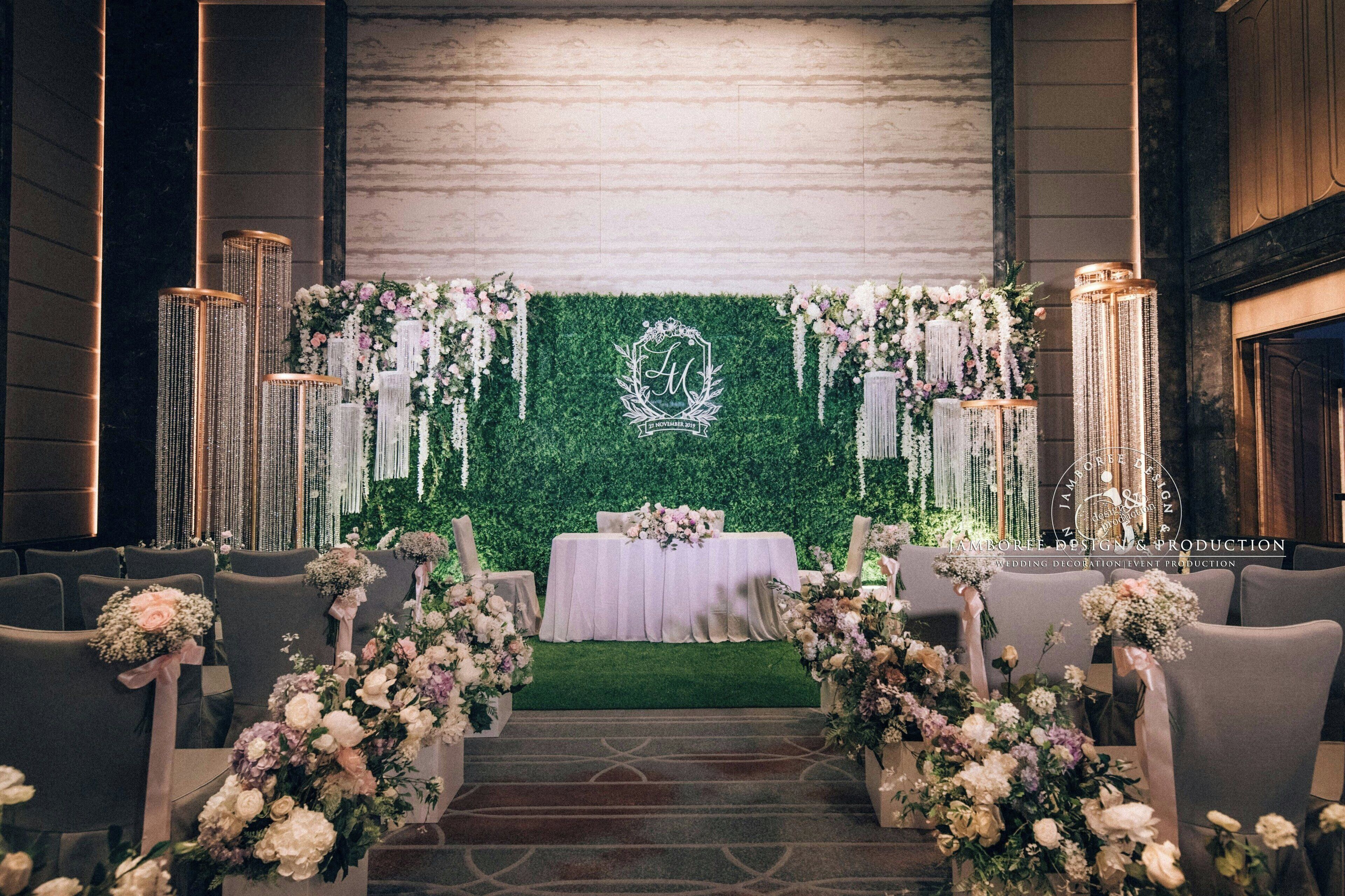 An indoor venue with a green wall backdrop, a white table with floral arrangements, and rows of chairs.