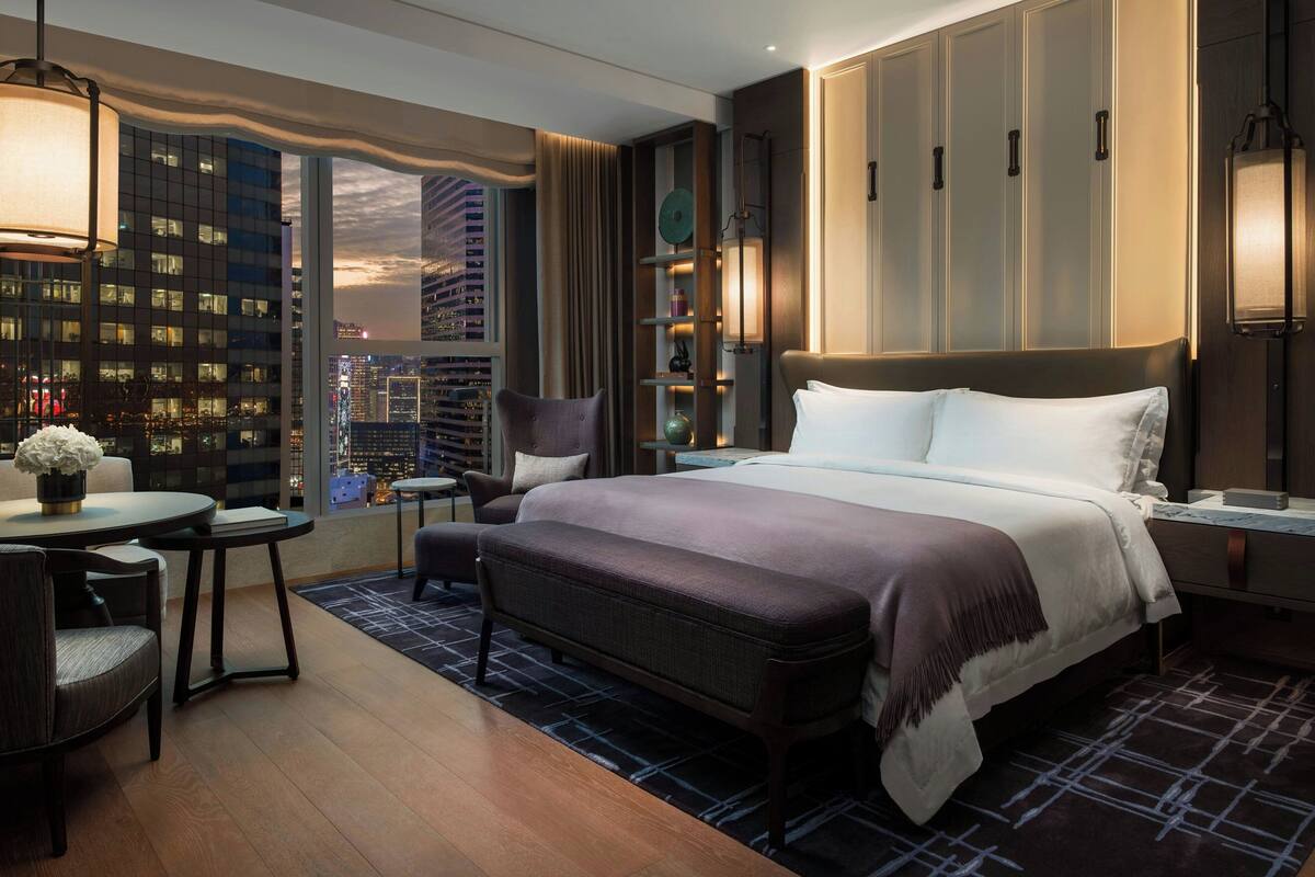 A hotel room with a large bed, a desk with a chair, a small table with a vase of flowers, and a view of the cityscape through a large window.