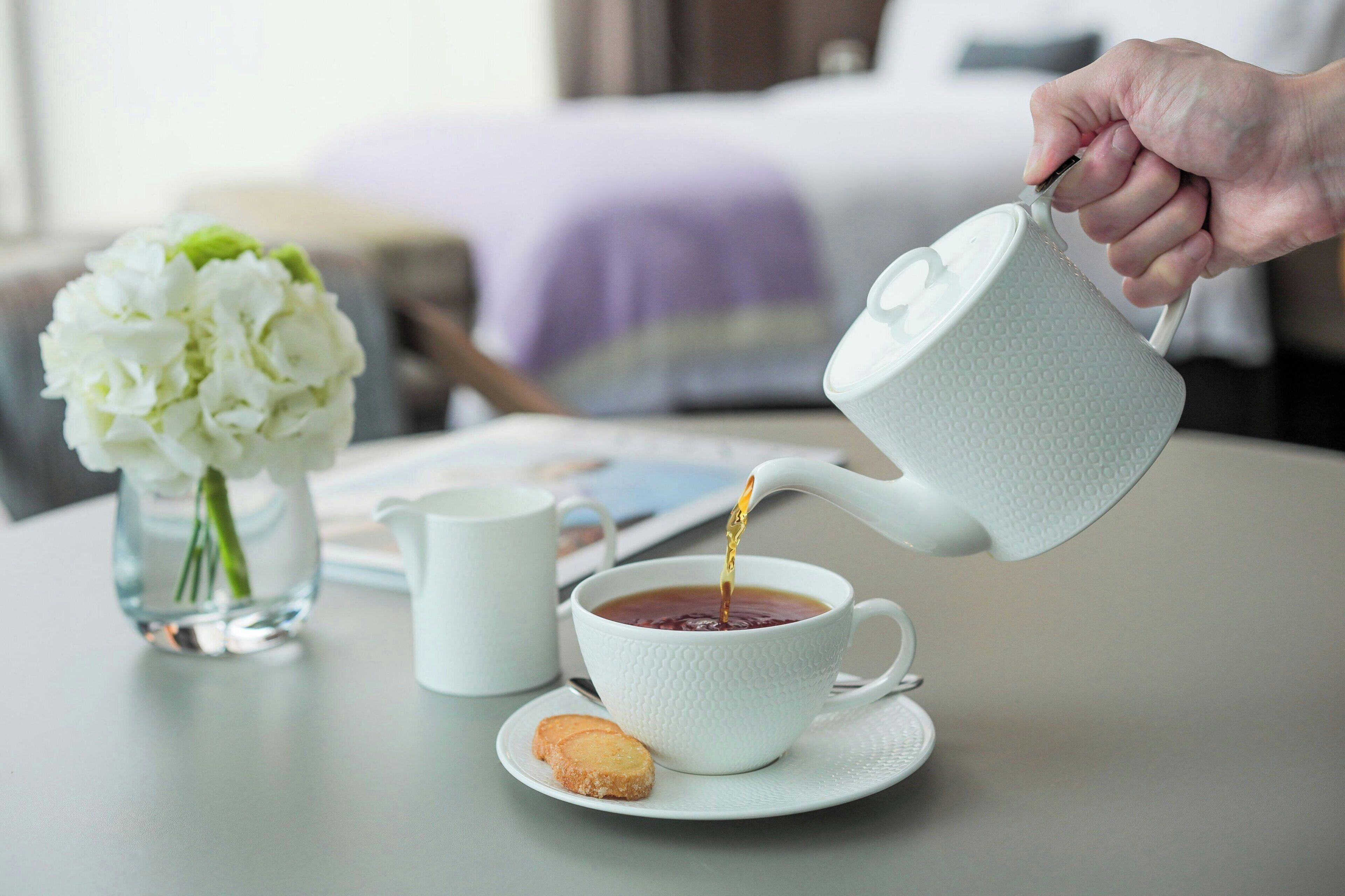 A hand pouring tea into a white cup on a table with a white teapot, a white teacup, a white saucer, and a white teapot.