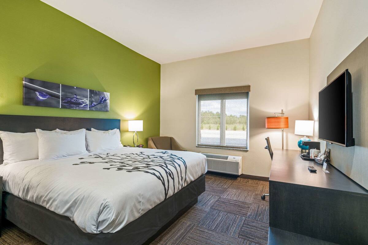 Ein Hotelzimmer mit einem großen Bett, einem Schreibtisch, einem Flachbildfernseher, einem Sessel, einem Fenster mit Ausblick und Kunstwerken an der Wand.