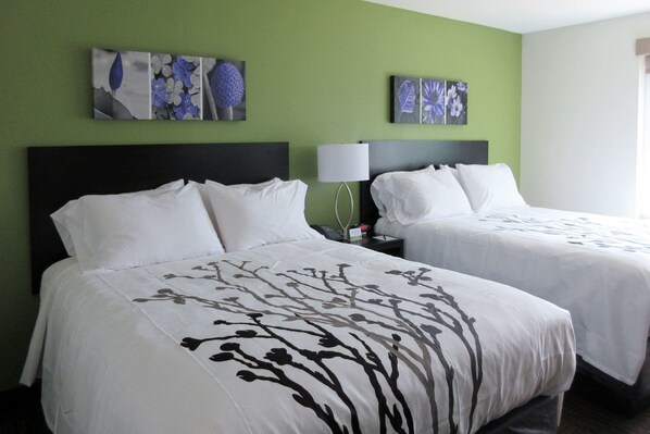 Two beds with white linens and a tree pattern, a nightstand with a lamp, and artwork on the green wall.