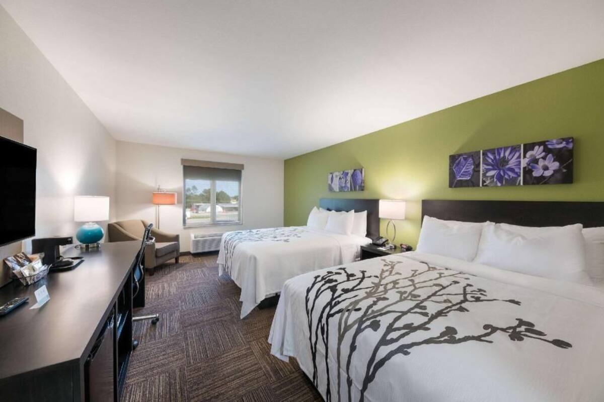 Ein Hotelzimmer mit zwei Betten, einem Schreibtisch, einem Fernseher, einem Fenster und Bildern an der Wand.