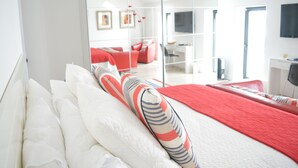 A modern hotel room with a bed, a red and white striped pillow, a white headboard, a red throw blanket, a large mirror, a desk, a chair, and a TV mounted on the wall. - Pier39Portrush (Portrush)