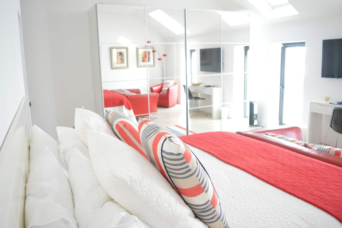 Une chambre d’hôtel moderne avec un lit, un oreiller à rayures rouges et blanches, une tête de lit blanche, un plaid rouge, un grand miroir, un bureau, une chaise et un téléviseur fixé au mur.