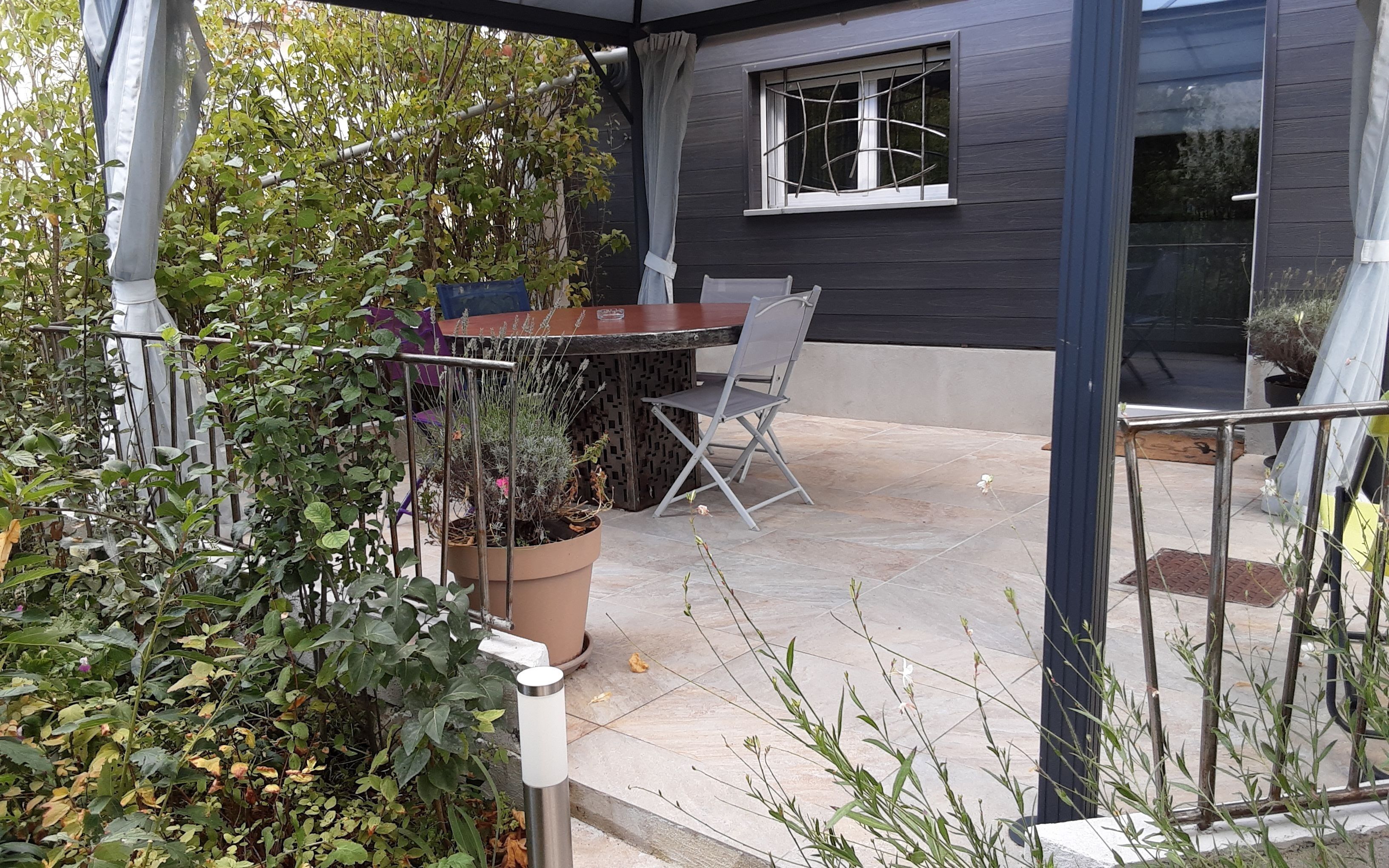 Une terrasse extérieure avec une table et des chaises, entourée de plantes en pot et d’un jardin.