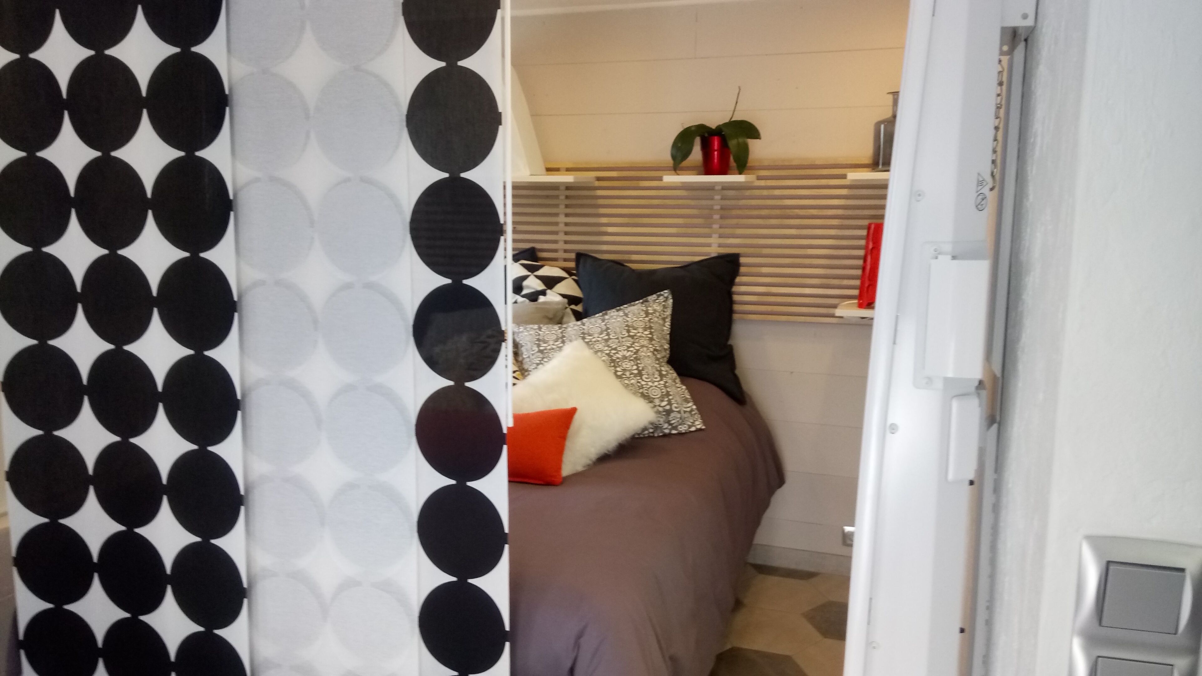 A bedroom with a patterned wall, a bed with multiple pillows, a window with blinds, and a small plant on the windowsill.