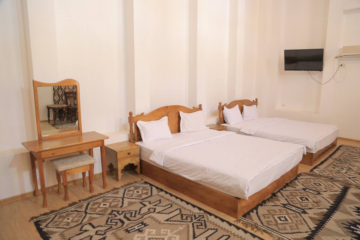A hotel room with two beds, a wooden desk, a chair, a television, and a patterned rug.