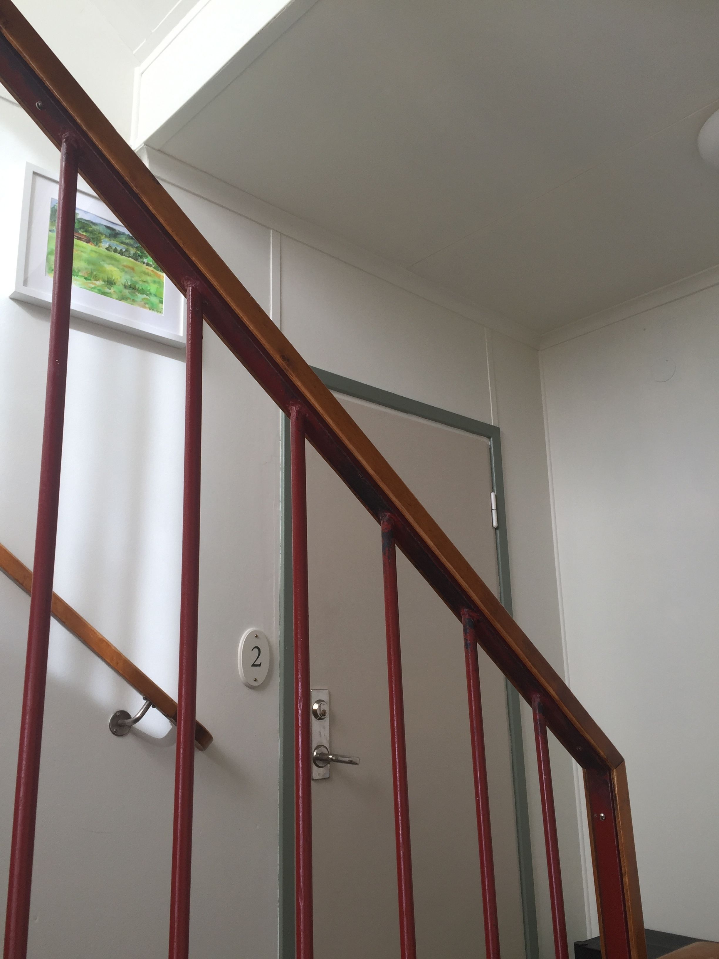 A staircase with a wooden handrail and red vertical supports, leading to a door with a number plate.