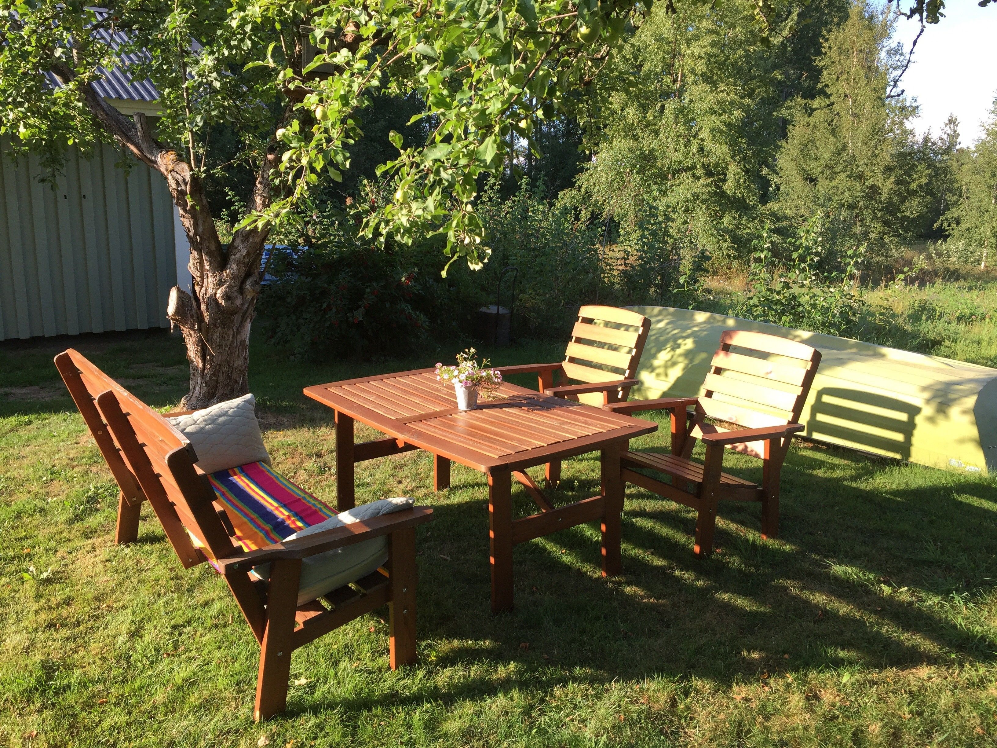 Een terras met een houten tafel en stoelen, een kleurrijk kussen en een boom die schaduw biedt.