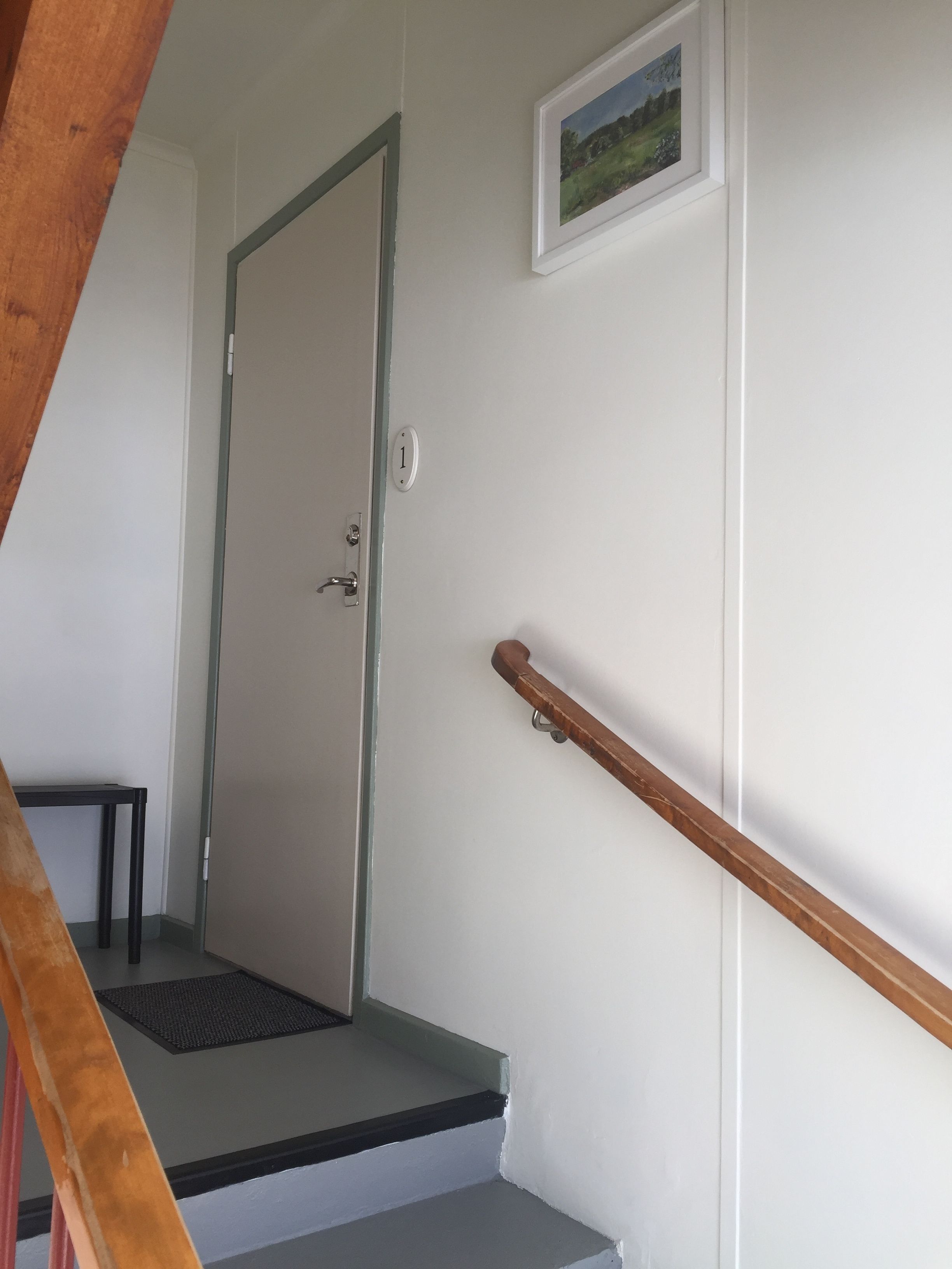 Una escalera con barandilla de madera, una puerta con placa de número y un pequeño cuadro enmarcado en la pared.