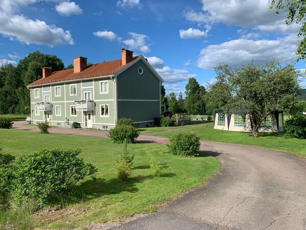 Una casa grande de dos pisos, con fachada verde y tejado rojo, rodeada de un césped cuidado y árboles.