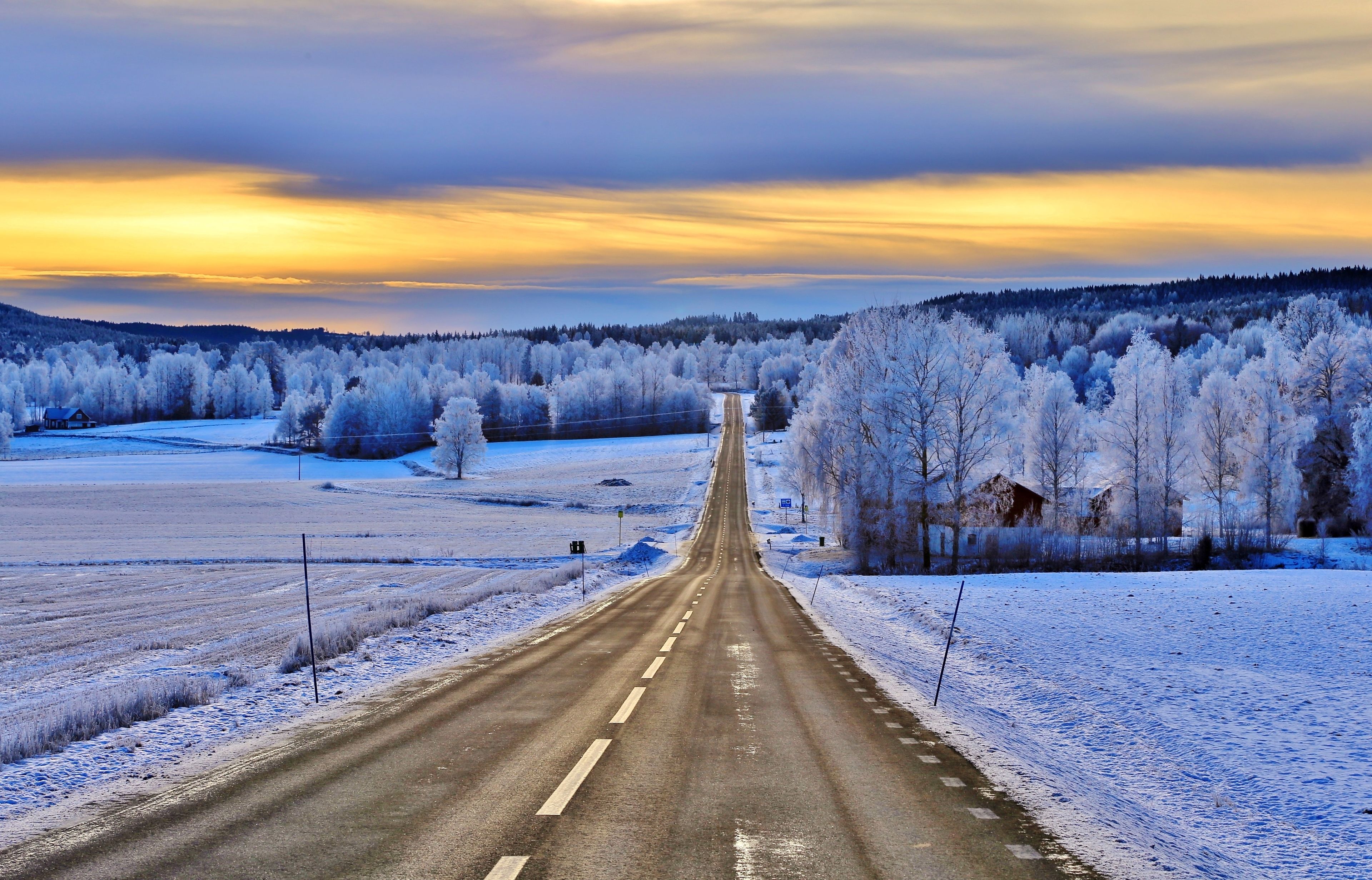 Une route enneigée qui traverse un paysage rural, avec des arbres et une maison visible au loin.