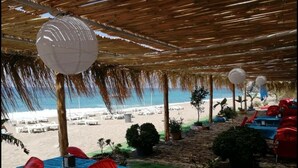 Un restaurante frente a la playa con terraza, techo de paja y vistas al mar.