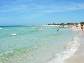 A beach with clear turquoise water, people swimming and walking along the shore, and a distant shoreline with buildings and trees. - VILLETTA MAGNOLIE in residence 400 mt from the sea, wi-fi free parking space. (San Vito Lo Capo)