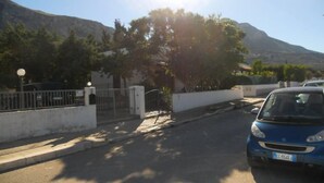 A street with a blue car parked on the right, a gated entrance, and a mountain in the background. - VILLETTA MAGNOLIE in residence 400 mt from the sea, wi-fi free parking space. (San Vito Lo Capo)