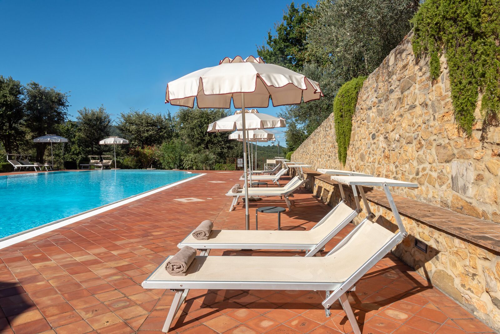 Un espace aménagé au bord de la piscine, avec des chaises longues et des parasols, un mur en pierre et une piscine à l’eau d’un bleu limpide.