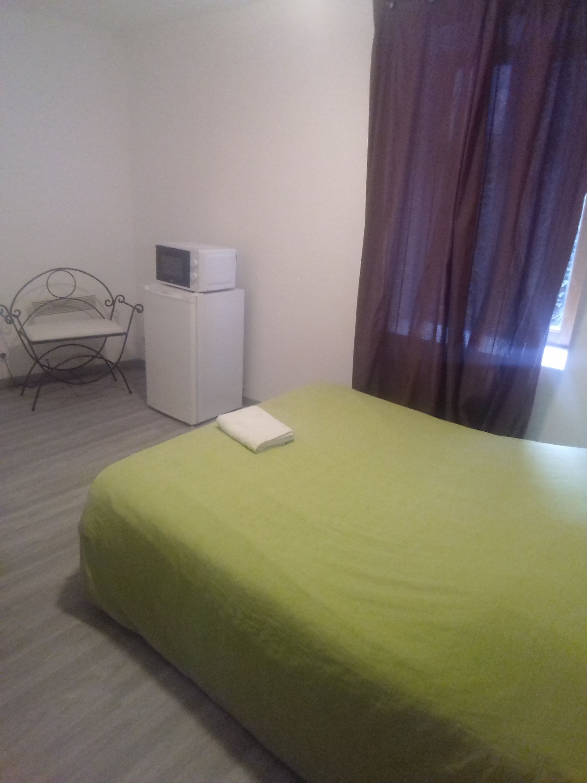 Una cama individual con una colcha verde, una mesita de noche blanca con un televisor, una silla de metal y una ventana con cortinas.