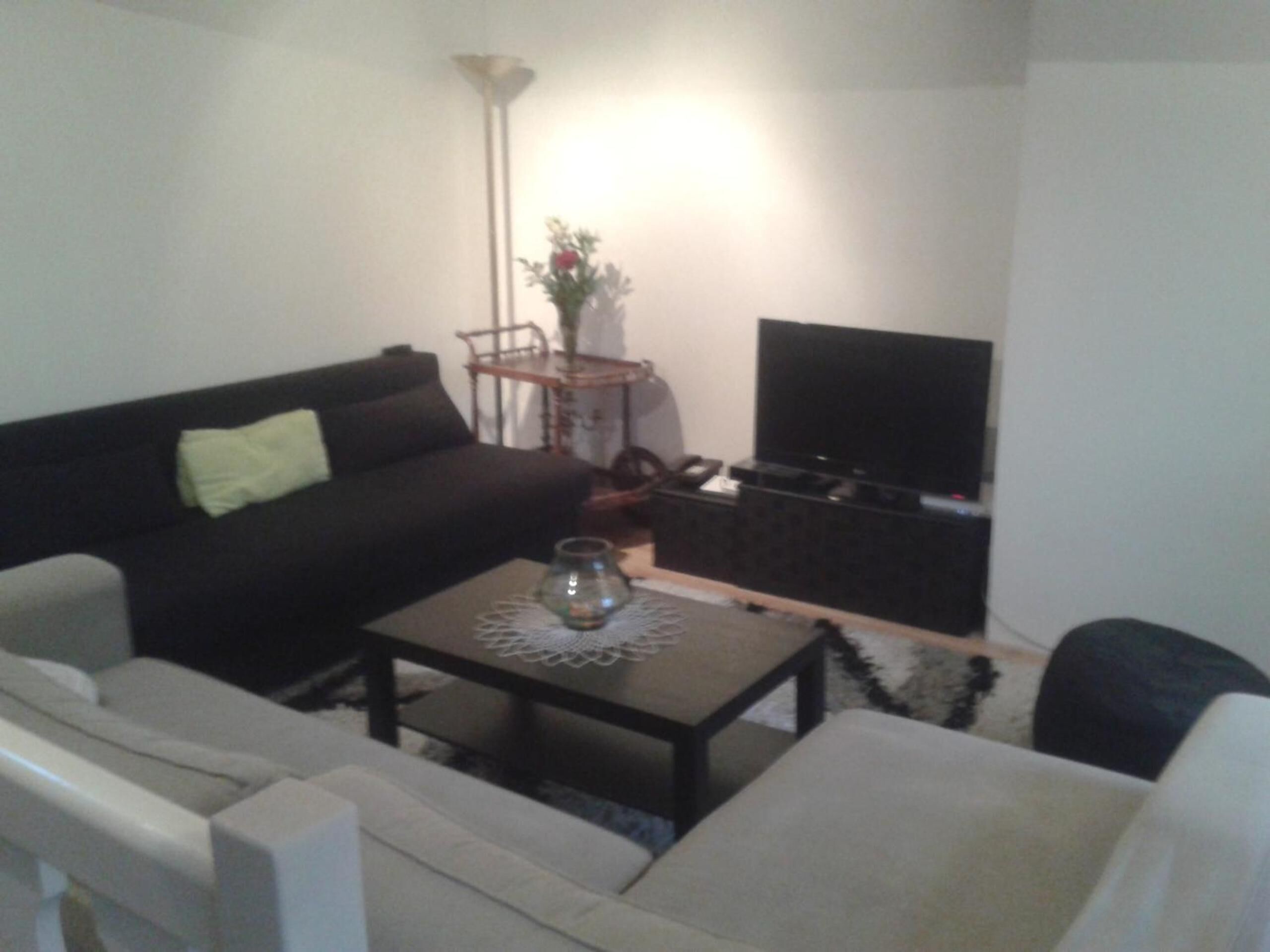 Una sala de estar con un sofá negro, un sillón blanco, una mesa de centro con un florero de vidrio y un televisor de pantalla plana sobre un soporte.