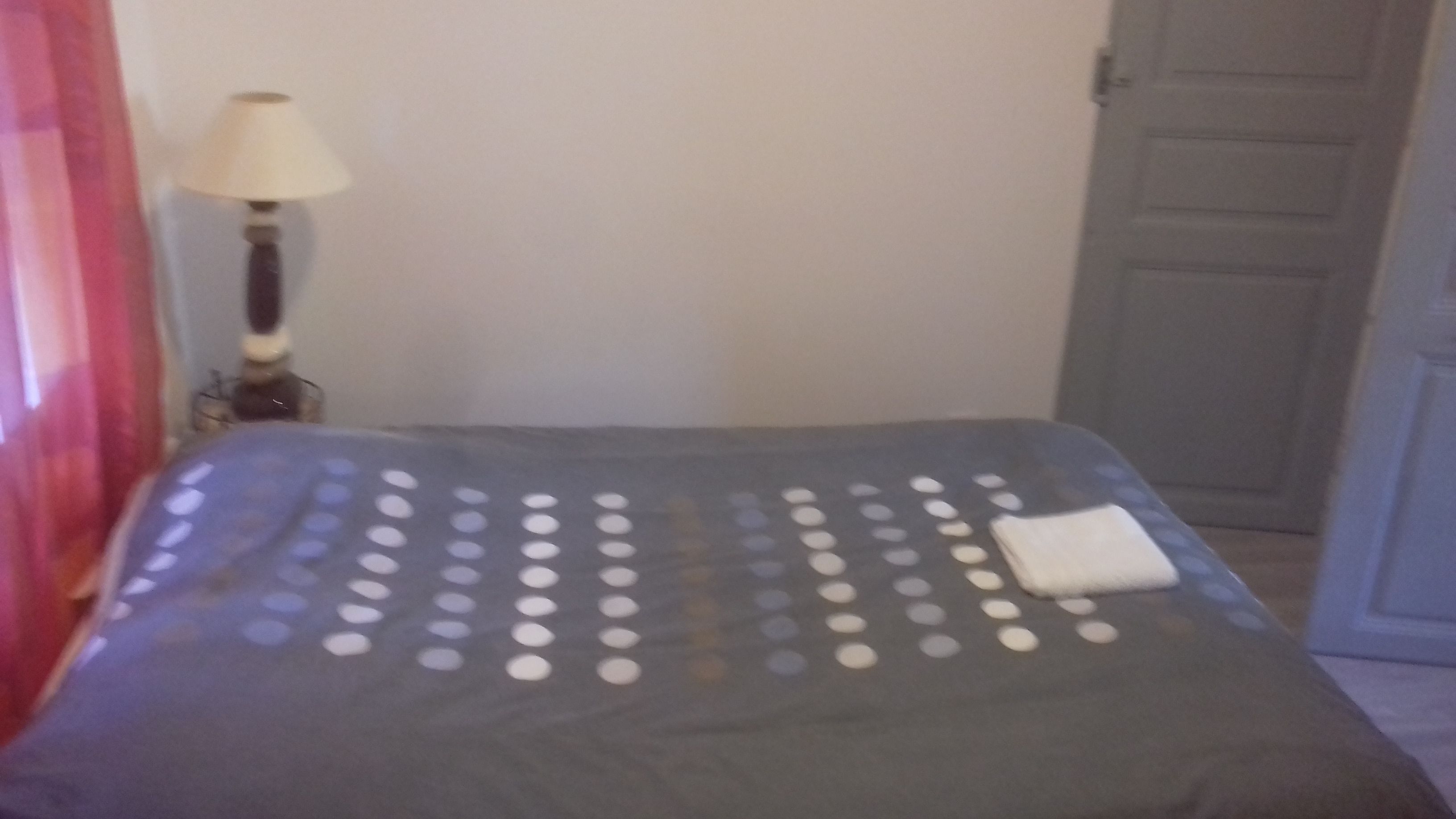 Ein Bett mit grauer Bettdecke und einem weißen Kopfkissen. Eine Lampe steht auf dem Nachttisch links.