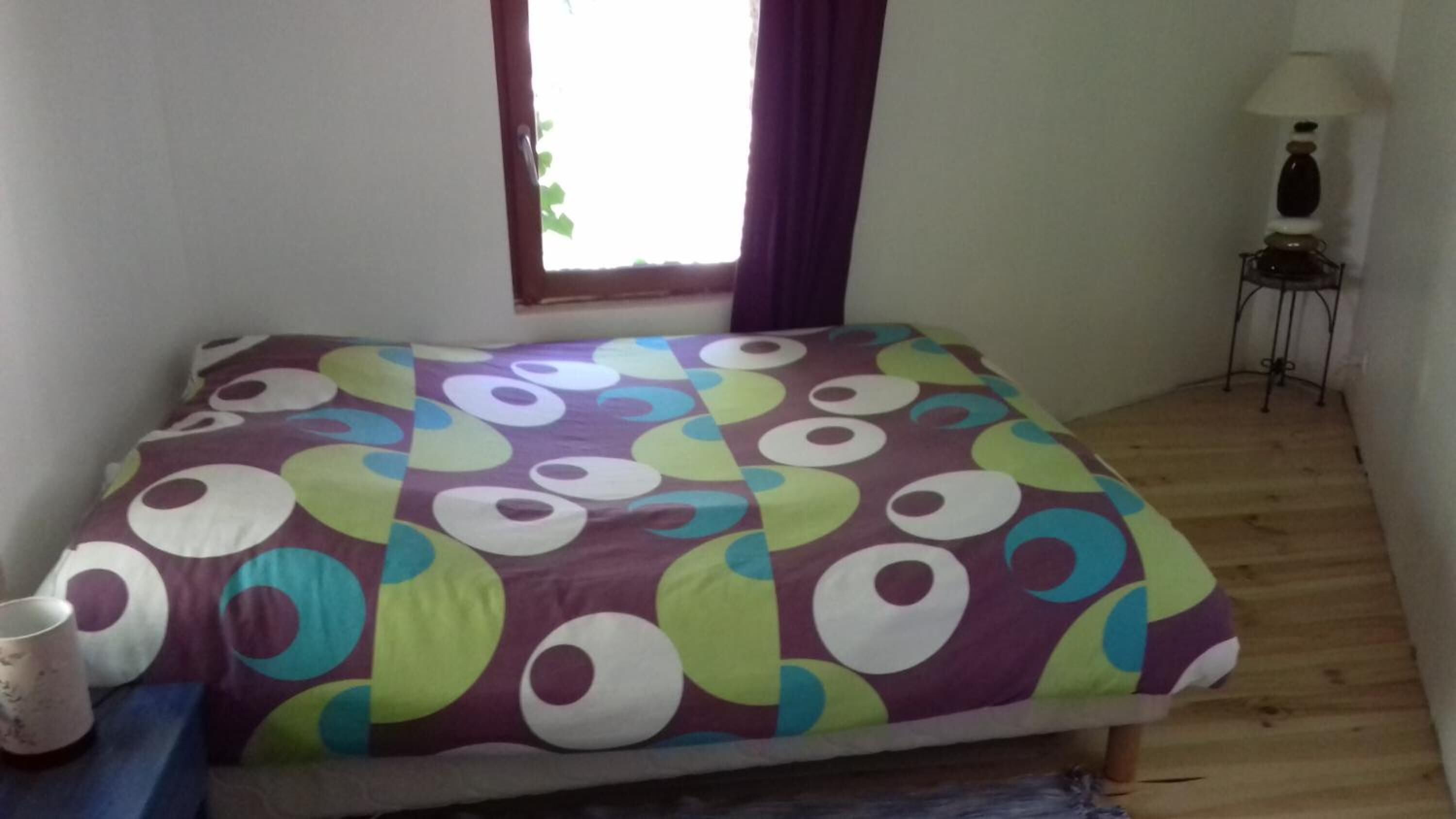 Quarto com cama coberta por um edredom colorido com bolinhas, janela com cortinas, mesinha com abajur e piso de madeira.