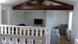 A room with a high ceiling, exposed wooden beams, a television on a stand, a small table, and a chair. - Le Trois Mâts (Thiers)