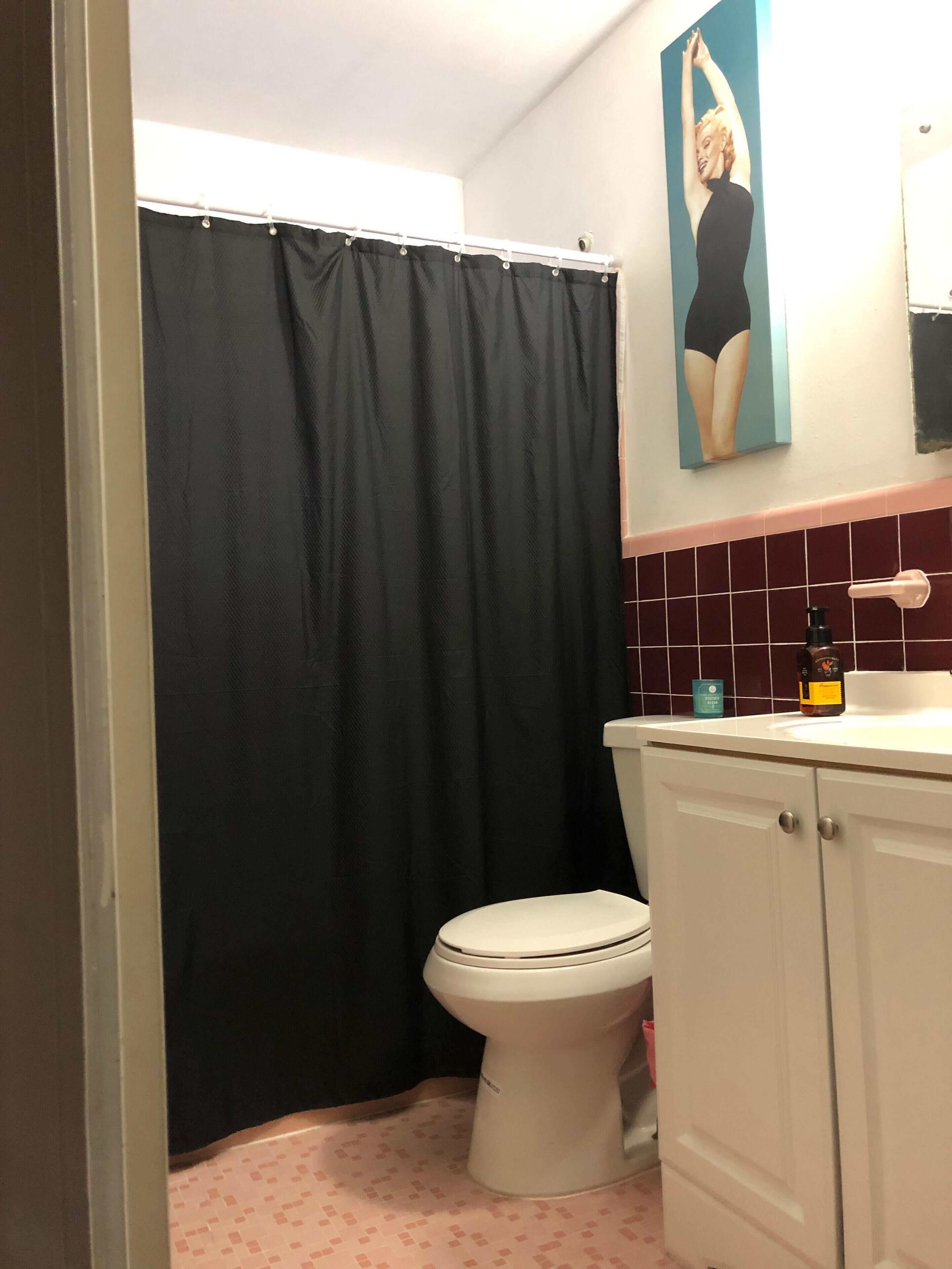 A bathroom with a toilet, a black shower curtain, a pink tiled floor, and a white sink with a soap dispenser.