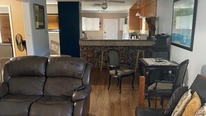 A living room with a brown leather sofa, a dark sofa with patterned cushions, and a wooden dining table with chairs. There is a kitchen area with a bar counter and a ceiling fan. - Relax in Galveston, Texas! (Galveston)