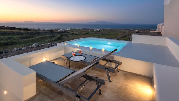 A rooftop pool with a table and chairs, surrounded by white walls, overlooking a coastal landscape at sunset. - Aplada Suites (Santorini)