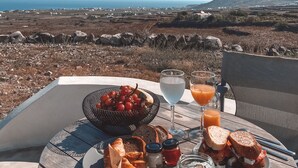 A table with a meal, including croissants, bread, and a sandwich, set against a scenic view of the ocean and hills. - Aplada Suites (Santorini)