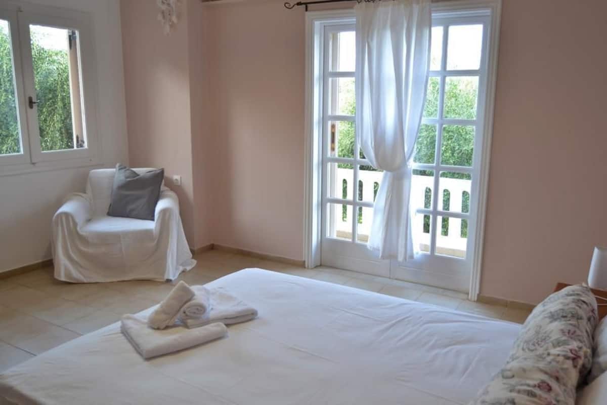 Une chambre avec un grand lit, une chaise, une fenêtre avec des rideaux et une vue sur la verdure.