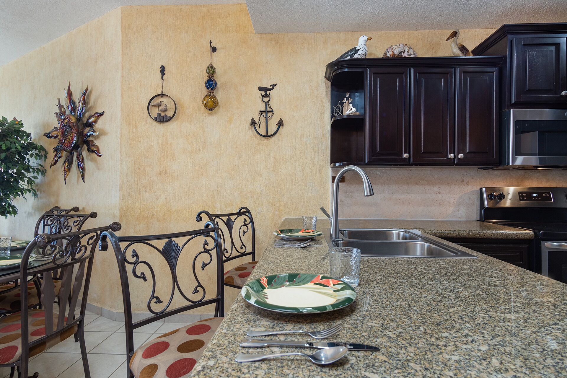 A kitchen with dark wooden cabinets, a granite countertop, a stainless steel sink, and a dining area with a metal chair and table.