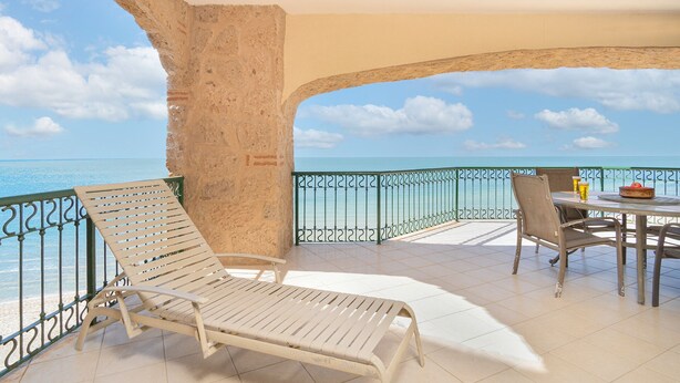A balcony with a white lounge chair, a dining table with chairs, and a view of the sea.