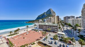 Paesaggio costiero con una montagna imponente, edifici fronte spiaggia e un’area di parcheggio.