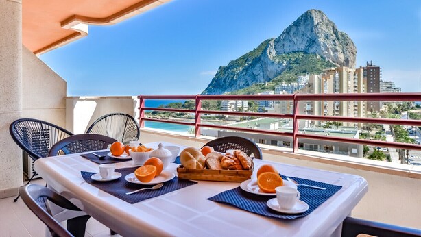 Un balcone con un tavolo apparecchiato per la colazione, affacciato su una città costiera con una montagna imponente.