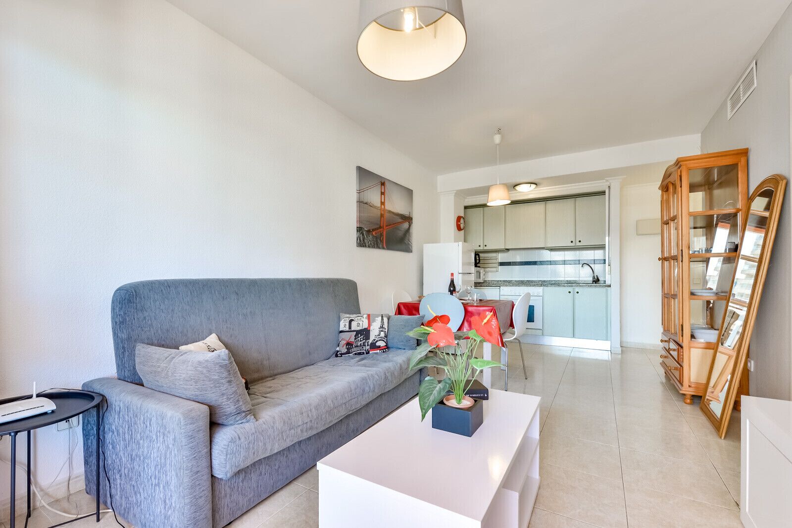 A modern living room with a grey sofa, a small table, and a potted plant. A kitchen is visible in the background with white cabinets and a dining area.
