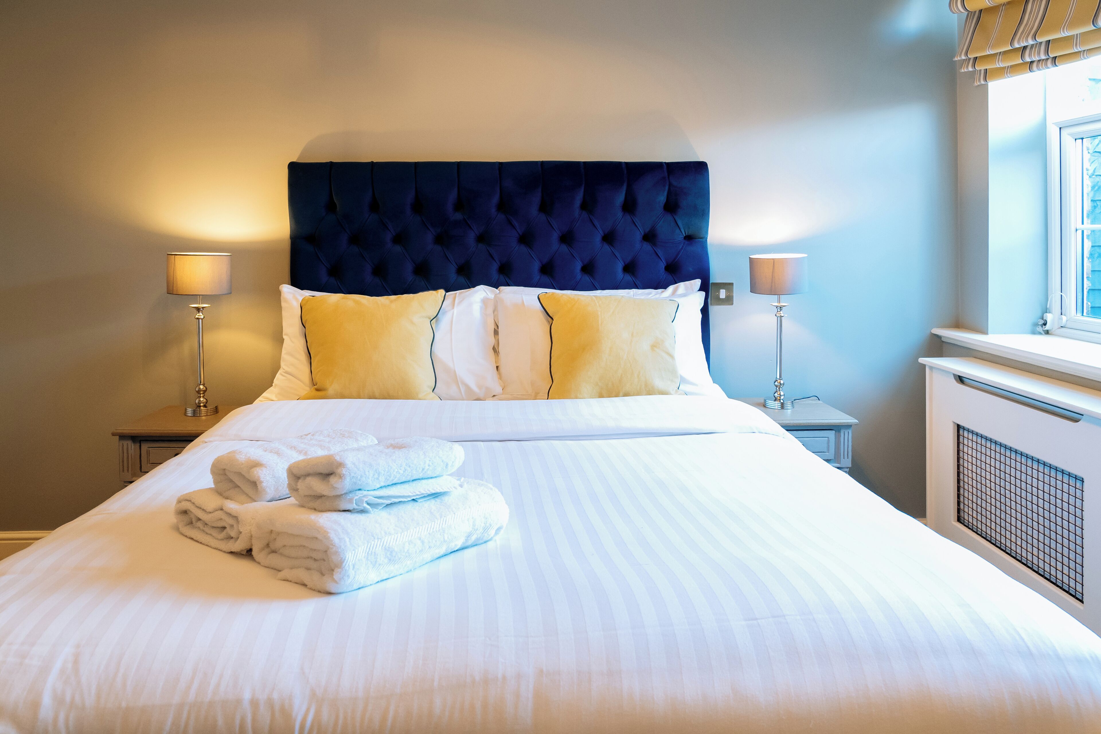 Een net opgemaakt bed met een gebonden hoofdbord, twee bedlampjes en een raam met jaloezieën.