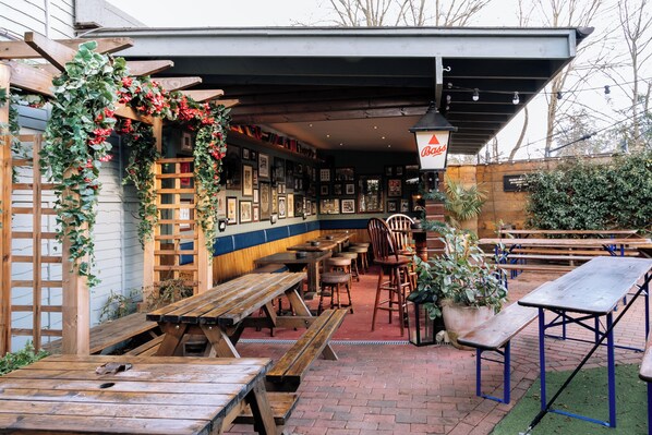 An outdoor patio area with wooden benches and tables, a covered seating area, and a variety of potted plants. - The Royal Oak (Woodford Green)