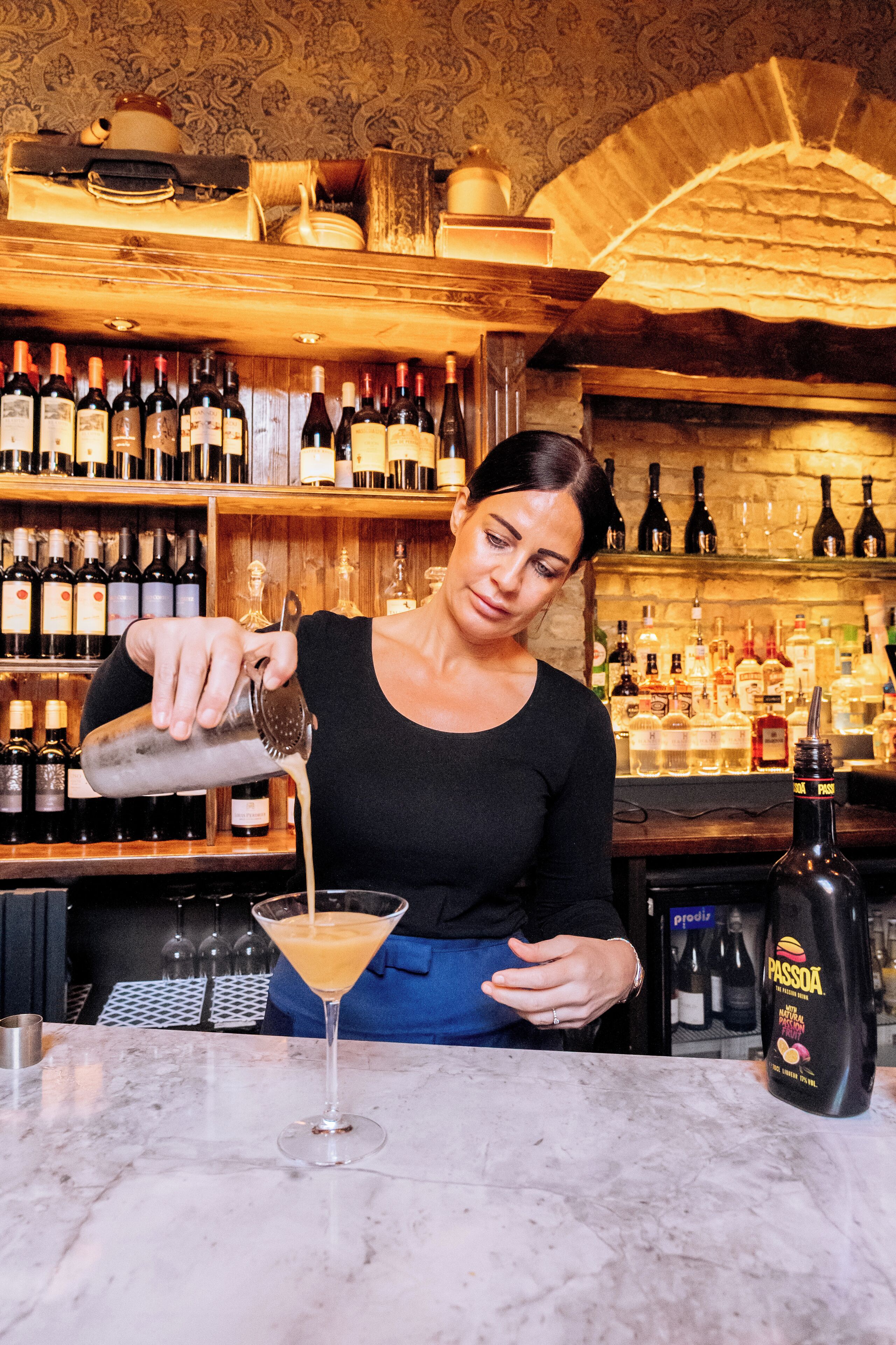 Un cantinero sirviendo una bebida en una copa de martini en un bar con estantes llenos de botellas al fondo.