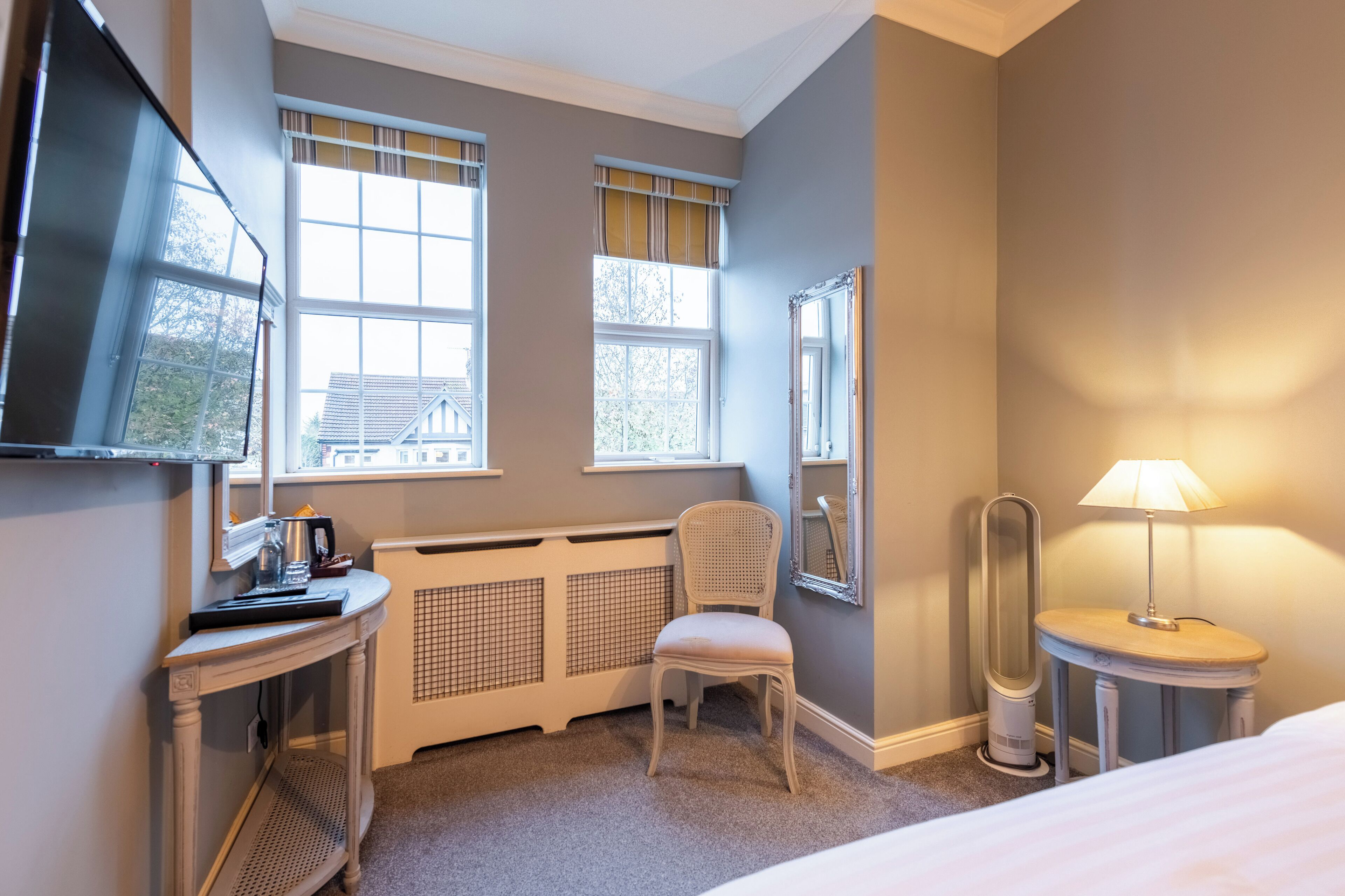 Habitación de hotel con una cama, un escritorio con lámpara, una silla, un radiador y dos ventanas con cortinas.