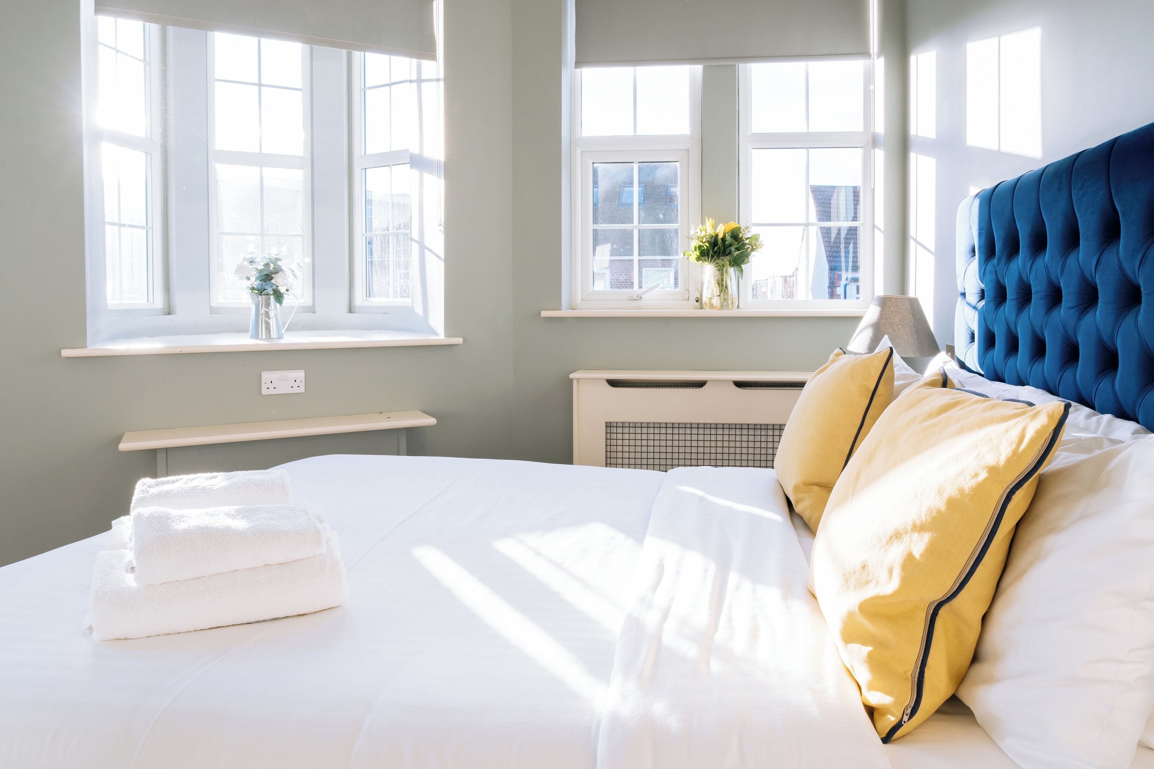 A neatly made bed with white linens, a blue headboard, and yellow pillows. There are two windows with white frames, one of which has a plant on the sill.