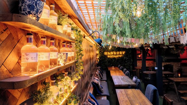 Een restaurant met houten planken waarop flessen staan, hangplanten en een neonbord waarop staat: "HANBURY DISTILLERY".