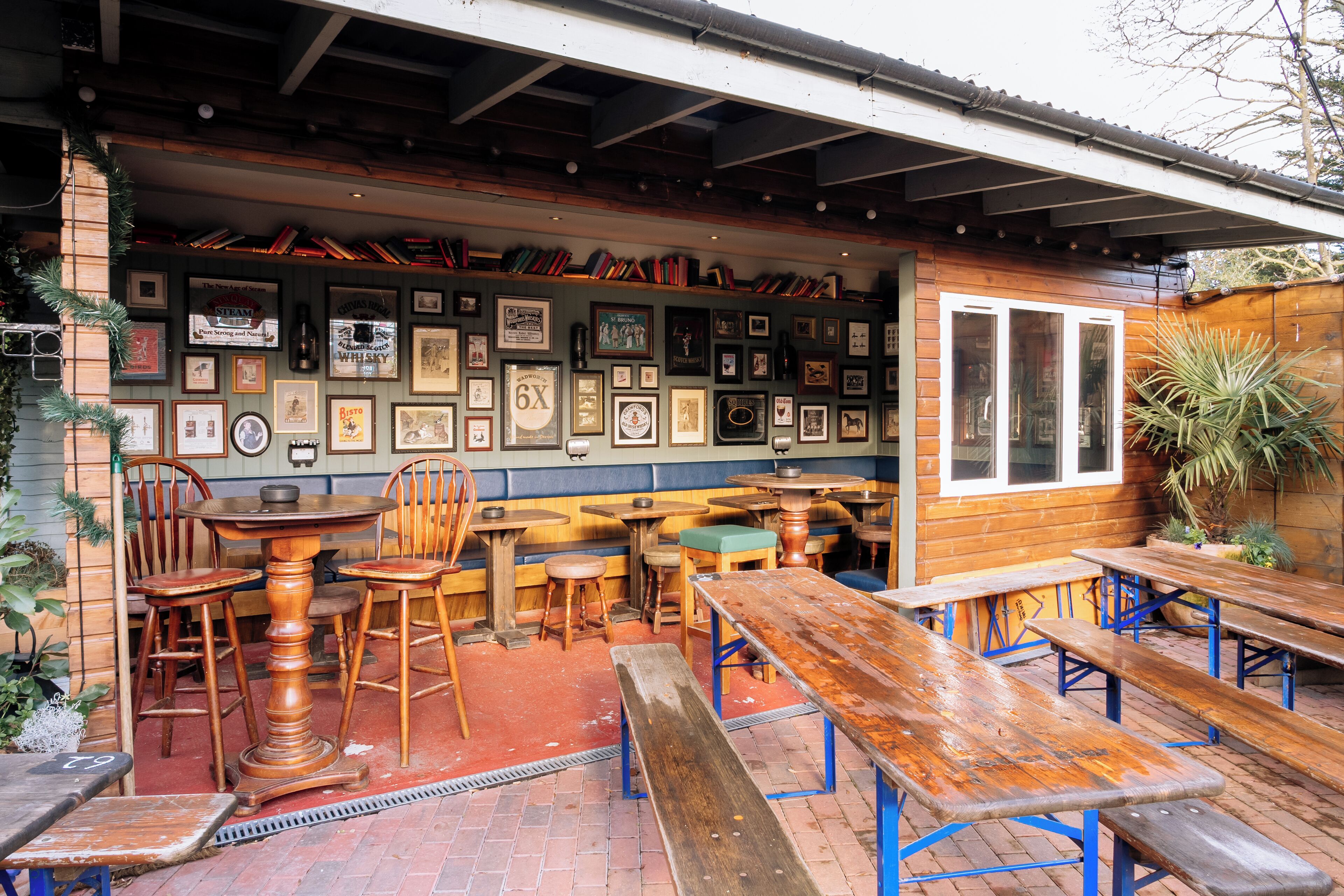 An outdoor patio with wooden tables and benches, surrounded by a brick floor and a wooden fence.