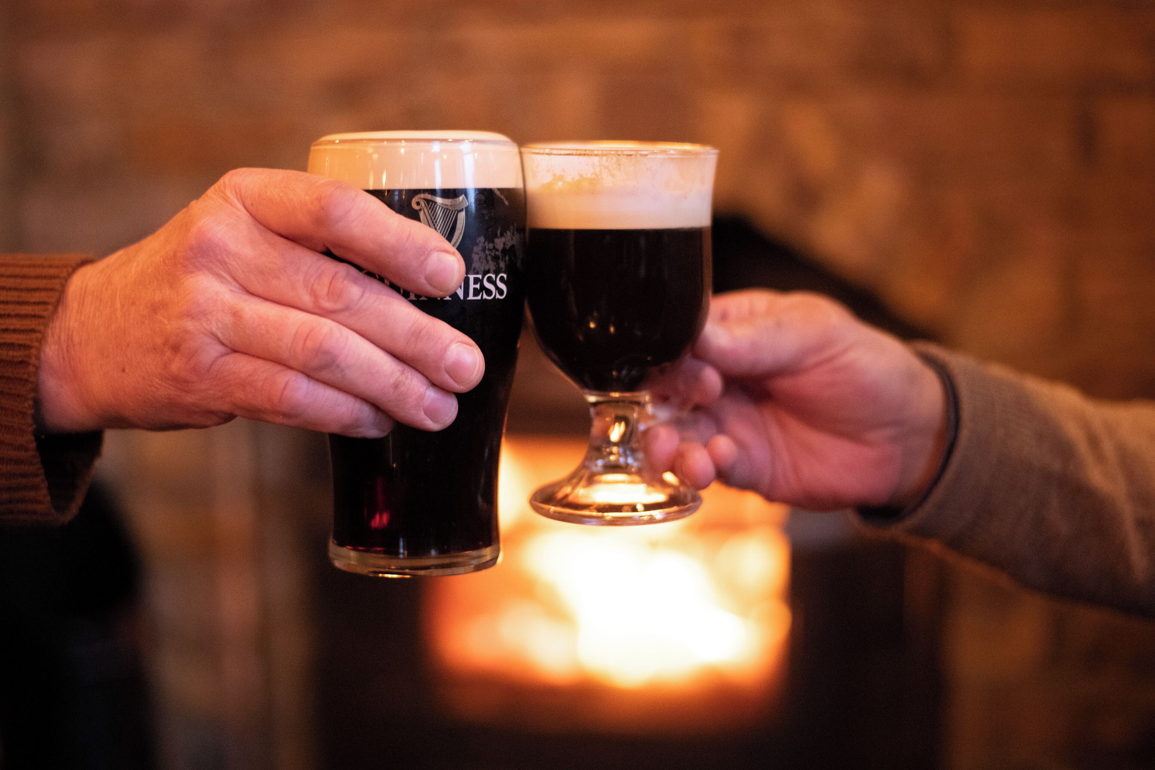 Two hands holding glasses of beer, one with a label and one without, in front of a fireplace.