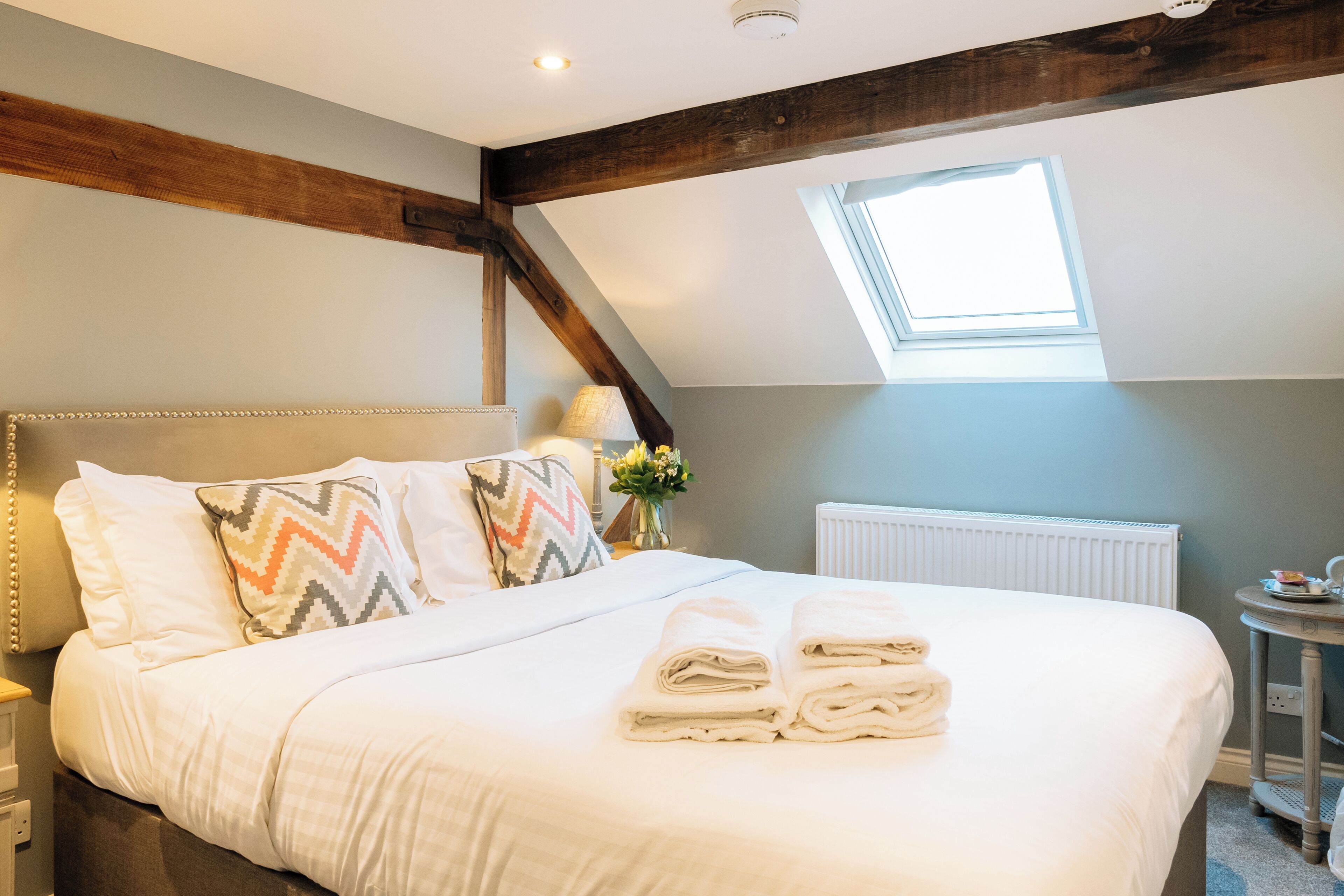A neatly made bed with pillows and towels, a small table with a lamp and flowers, and a skylight in the room.