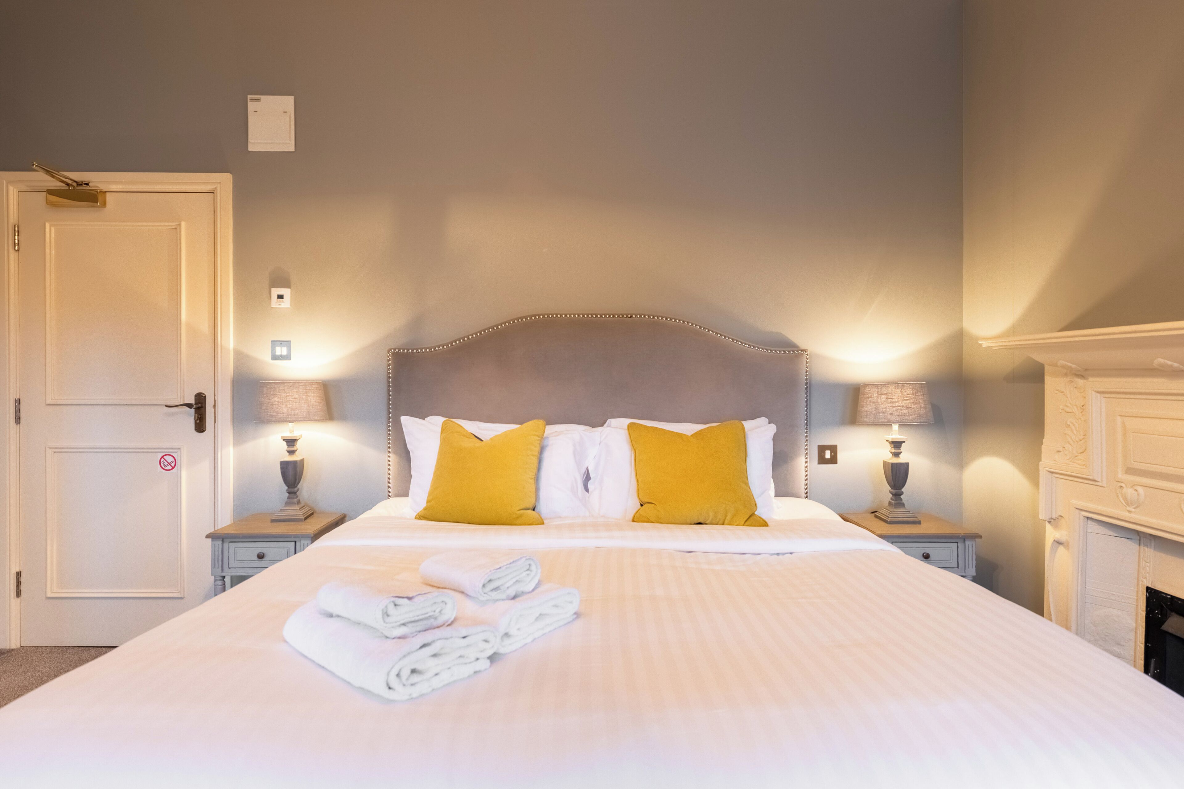A neatly made bed with white linens and two yellow pillows, a bedside table with a lamp, and a door with a "no entry" sign.