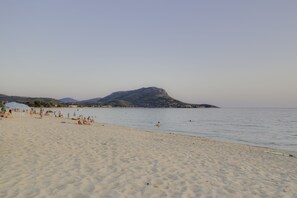 A beach with people sunbathing and swimming, a clear sky, and a mountain in the background.