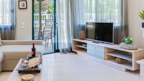 A modern living room with a sofa, a TV on a wooden stand, and a dining area visible through the glass doors. - Toroni Blue (Sithonia)