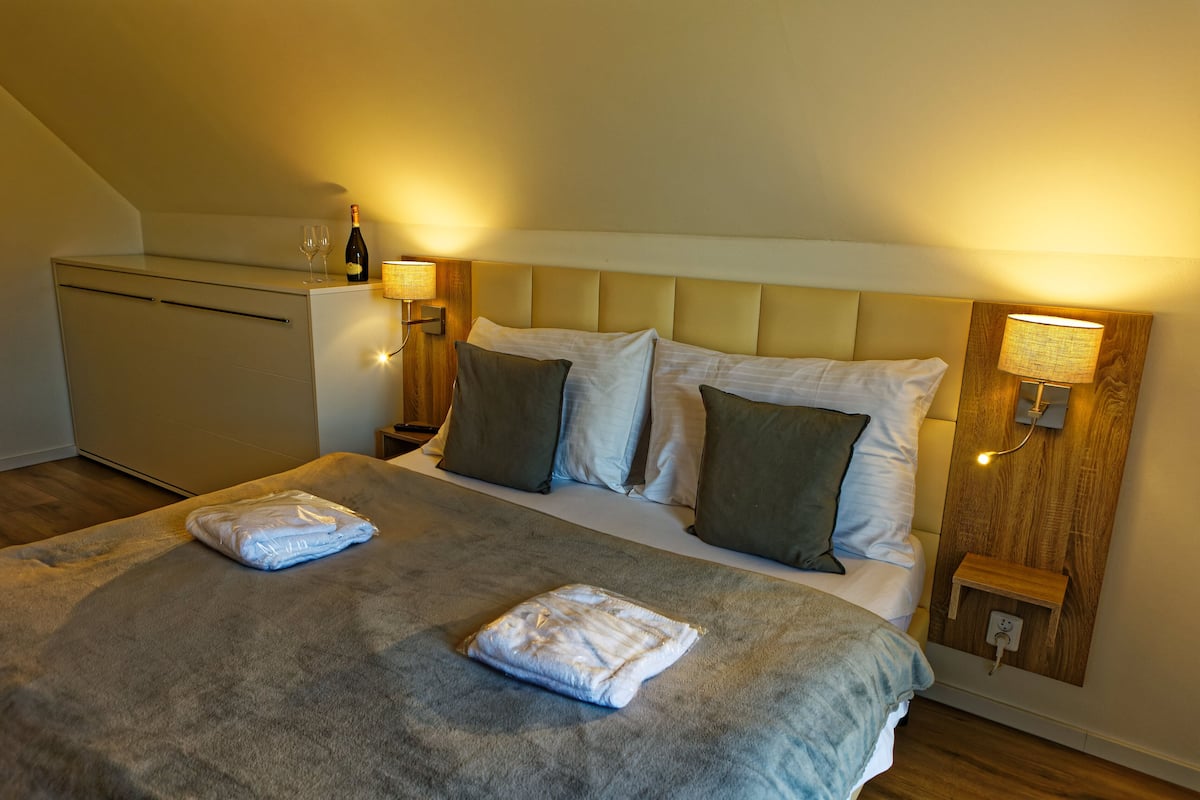 A neatly made bed with a grey blanket and two folded towels, a bedside table with a lamp, and a bottle of wine on the nightstand.